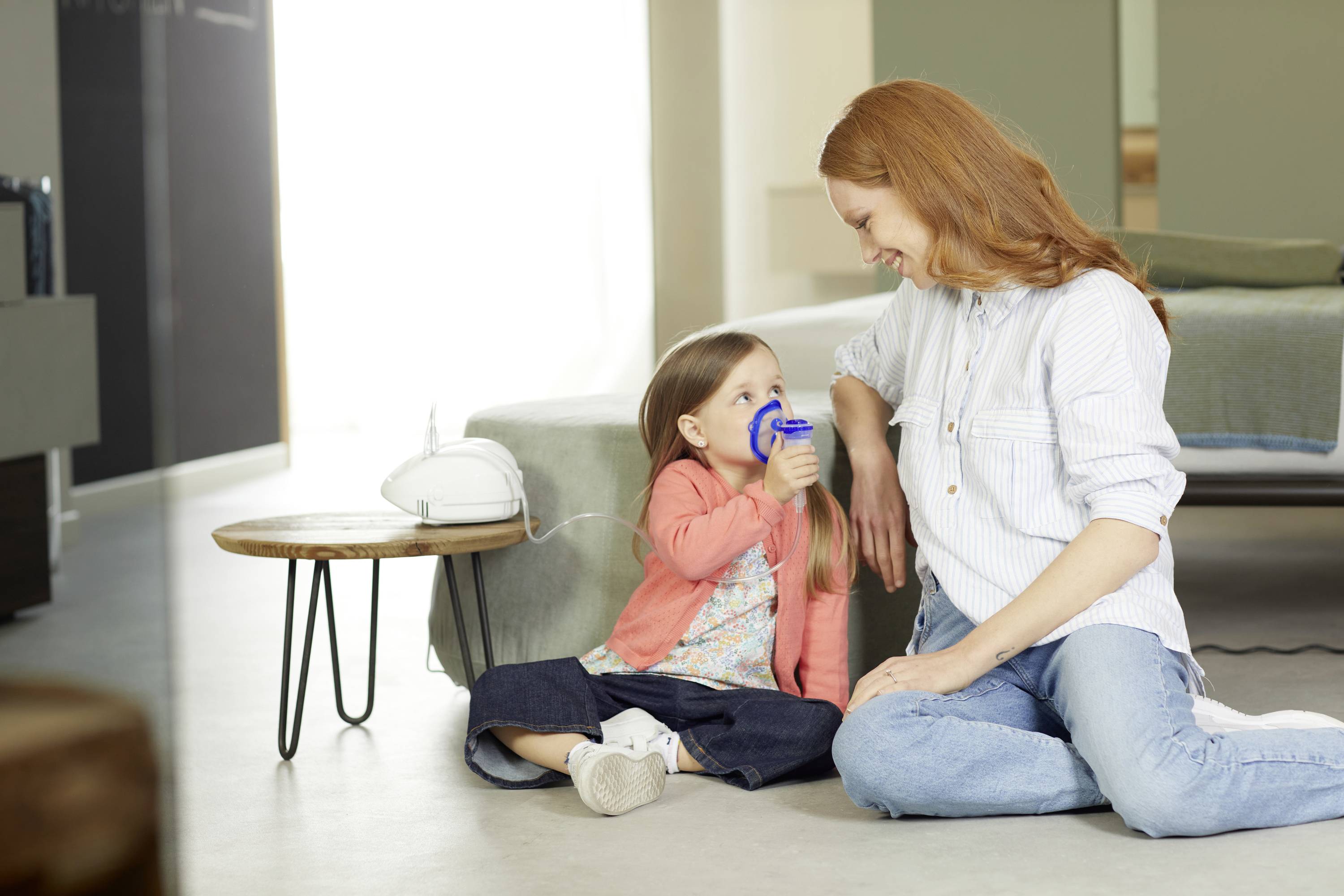 Mother and child on the floor. The child is using an inhaler. The mother looks on smiling. Living room in the background.<br><br>Note: The translation is already in British English and does not require modification.