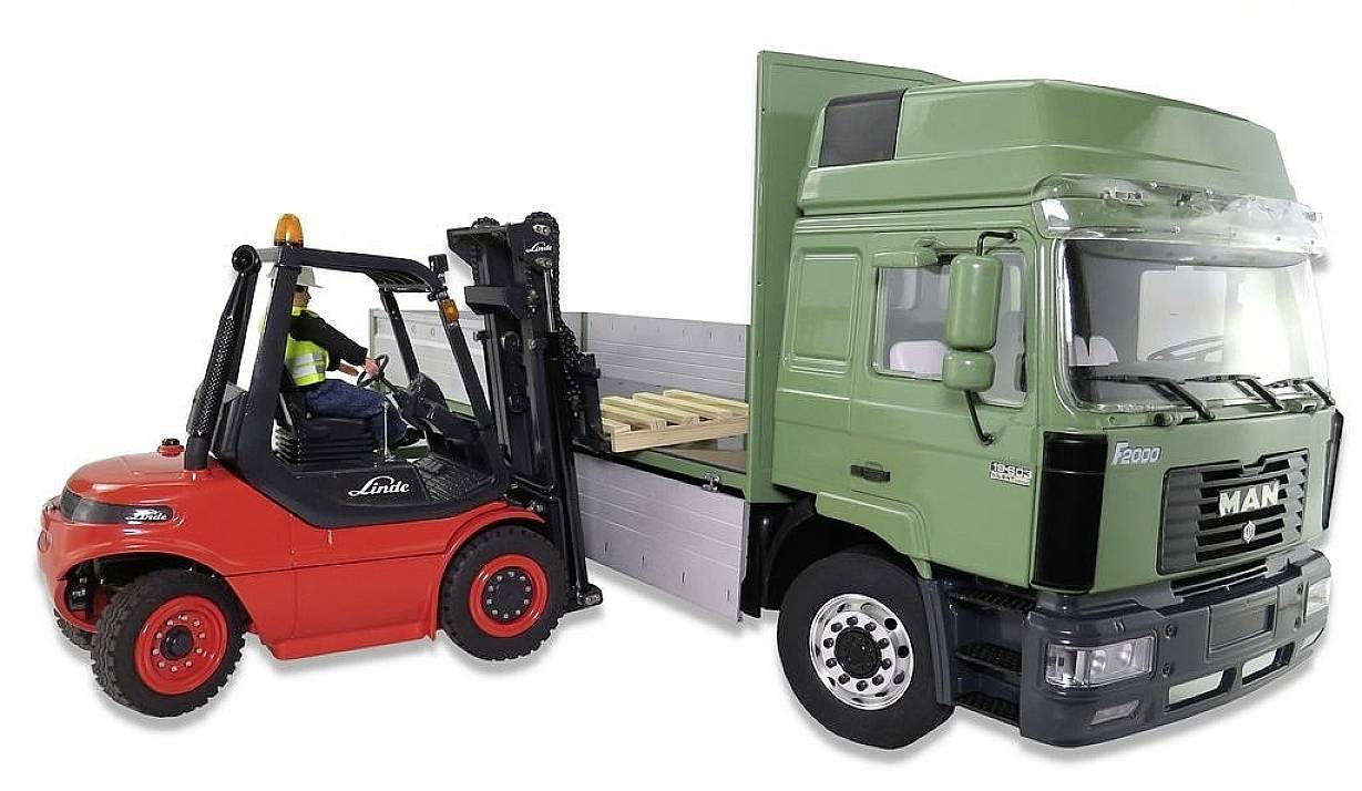 A red forklift is lifting a wooden pallet onto the loading area of a green lorry. A worker is seated in the forklift.