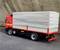 A red lorry with a grey tarpaulin is parked on an asphalted surface in front of a concrete wall.