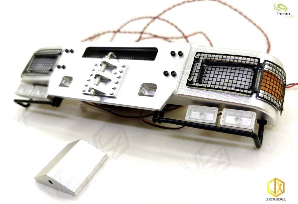A silver model component of a vehicle bumper with grilles, reflectors, and cables lies on a table.