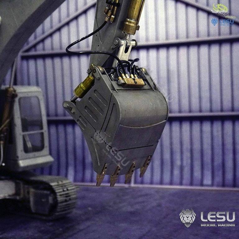 An excavator arm with a grab bucket inside a hall. The background shows metal structures. Brand logos are visible in the corner.