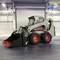 A grey mini loader with a red bucket attachment stands in an industrial hall. A large roller shutter door is visible in the background.