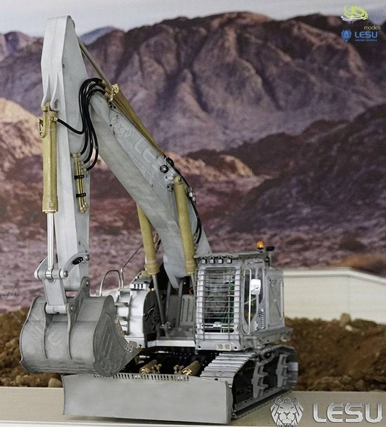 A silver excavator with a large bucket stands in front of a mountainous landscape. In the background, there are mounds of earth and rocky hills.