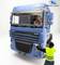 A large blue lorry with a driver inside. A person wearing a yellow high-visibility vest is standing in front of the lorry.