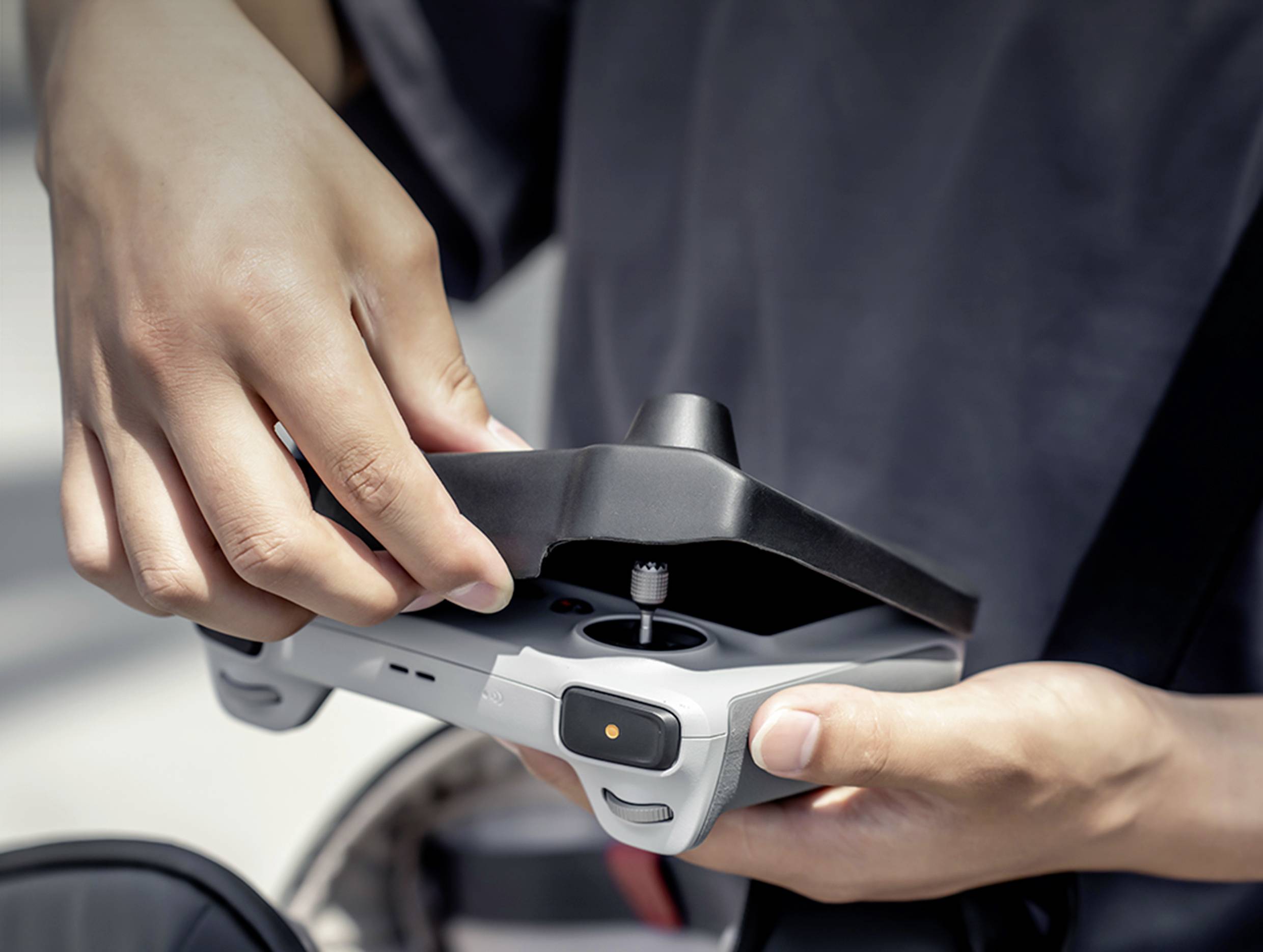A person is holding a drone remote control with both hands. Their left hand is adjusting the antenna. The background is out of focus.