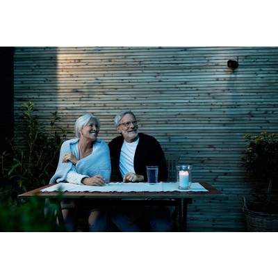 An elderly couple sits outdoors at a table, smiling and holding hands. A glass of water and a burning candle are on the table.