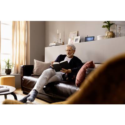 An elderly gentleman is sitting relaxed on a sofa, reading a book. Shelves with decorations and plants are visible in the background.