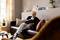 An elderly gentleman is sitting relaxed on a sofa, reading a book. Shelves with decorations and plants are visible in the background.