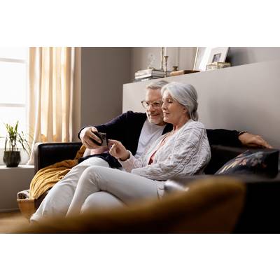 An older couple sit on a sofa, looking together at a smartphone and smiling. In the background, books and a bunch of plants are visible.