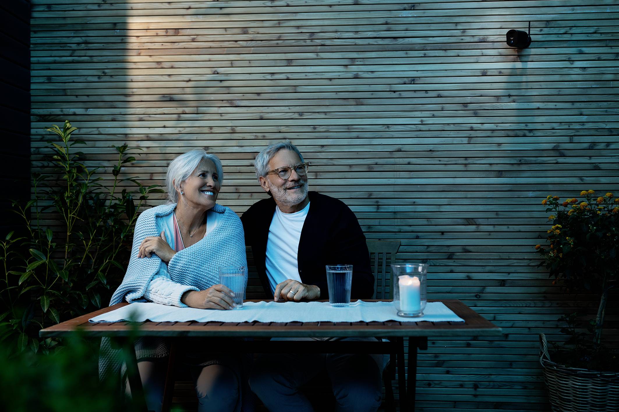 An older couple sits at a wooden table outdoors, surrounded by plants. They look relaxed and are smiling, with a candle burning on the table.
