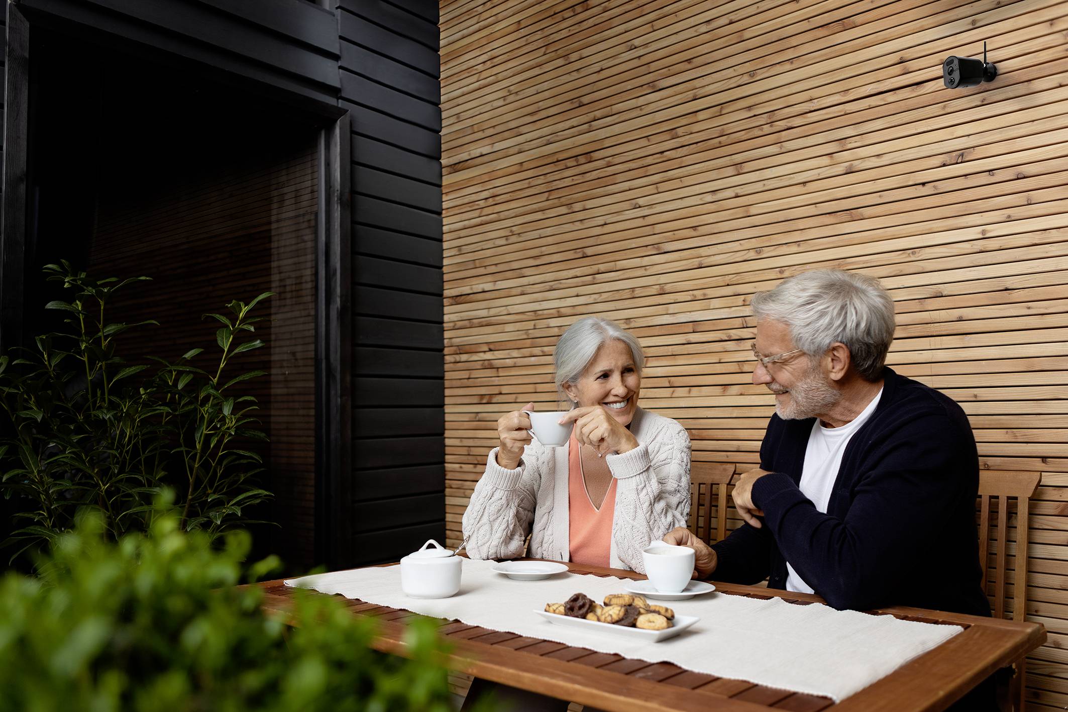 An older couple sit at a table on a terrace, drinking tea. A teapot and biscuits are placed before them. They smile and chat with one another.