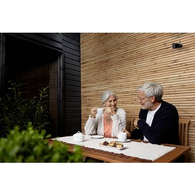 An older couple sit at a table on a terrace, drinking tea. A teapot and biscuits are placed before them. They smile and chat with one another.