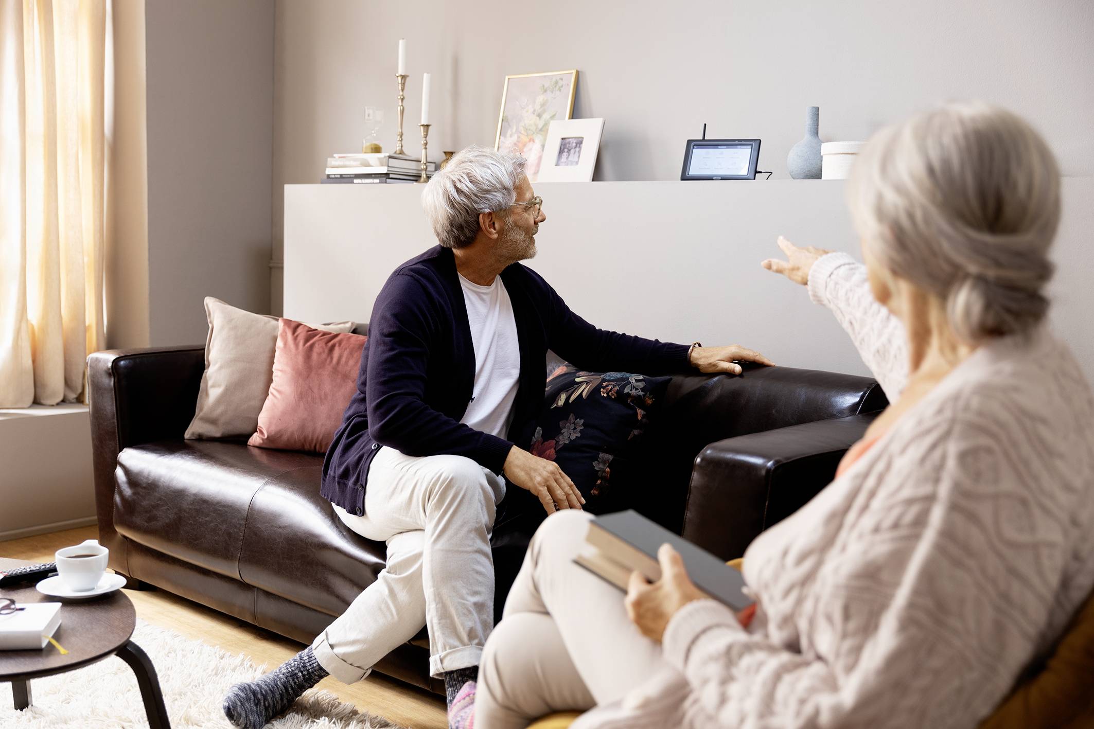 Two older people are sitting in the living room on sofas. One person is pointing at an electronic device on a shelf.