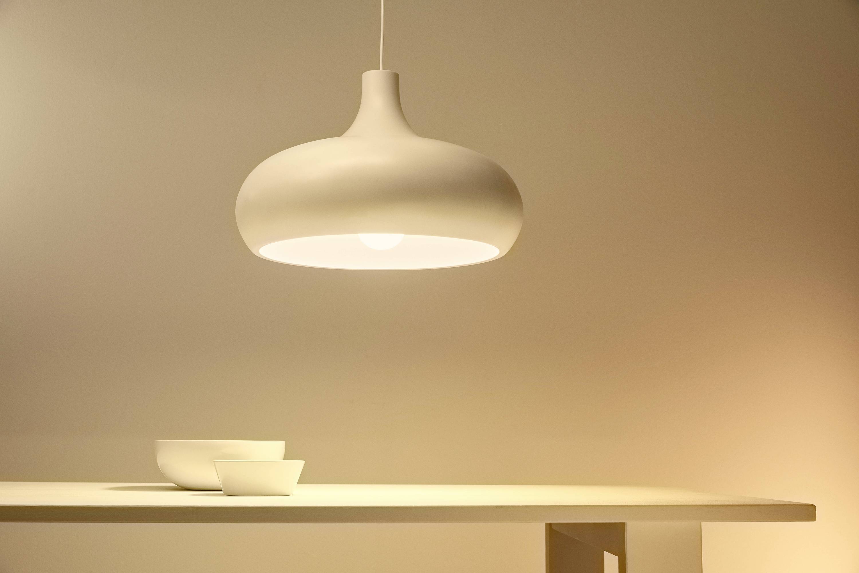 A white, modern pendant light illuminates a table with two white bowls positioned on one side. Calm, minimalist atmosphere.