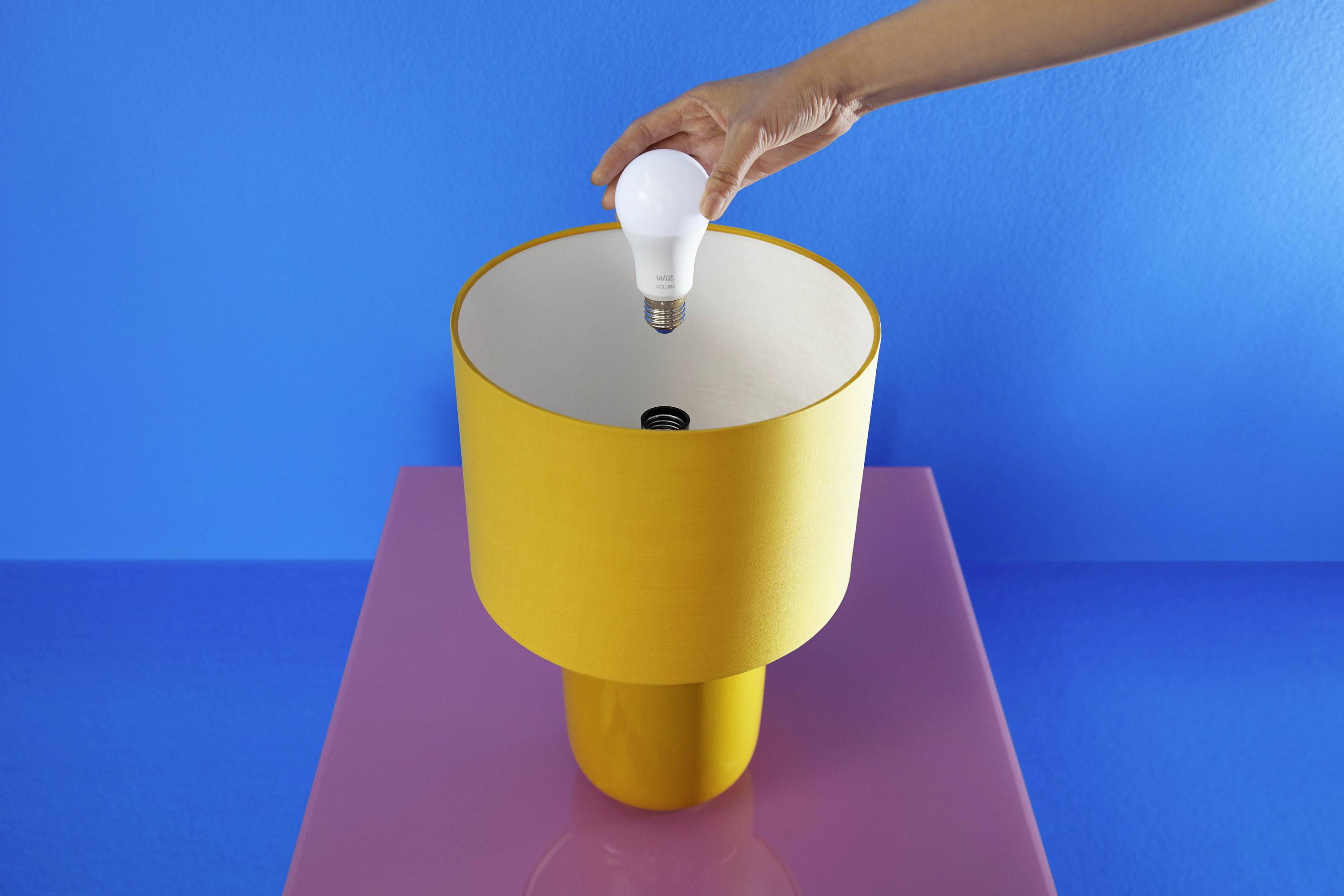 A hand changes a light bulb in a yellow table lamp on a pink table against a blue background.