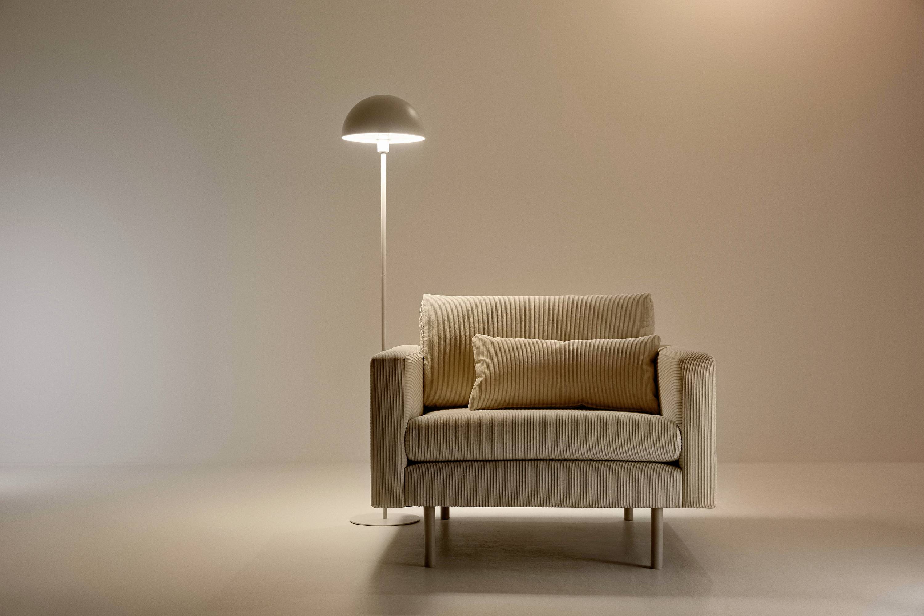 A beige armchair with cushions sits beside a modern standard lamp in a minimalist room.