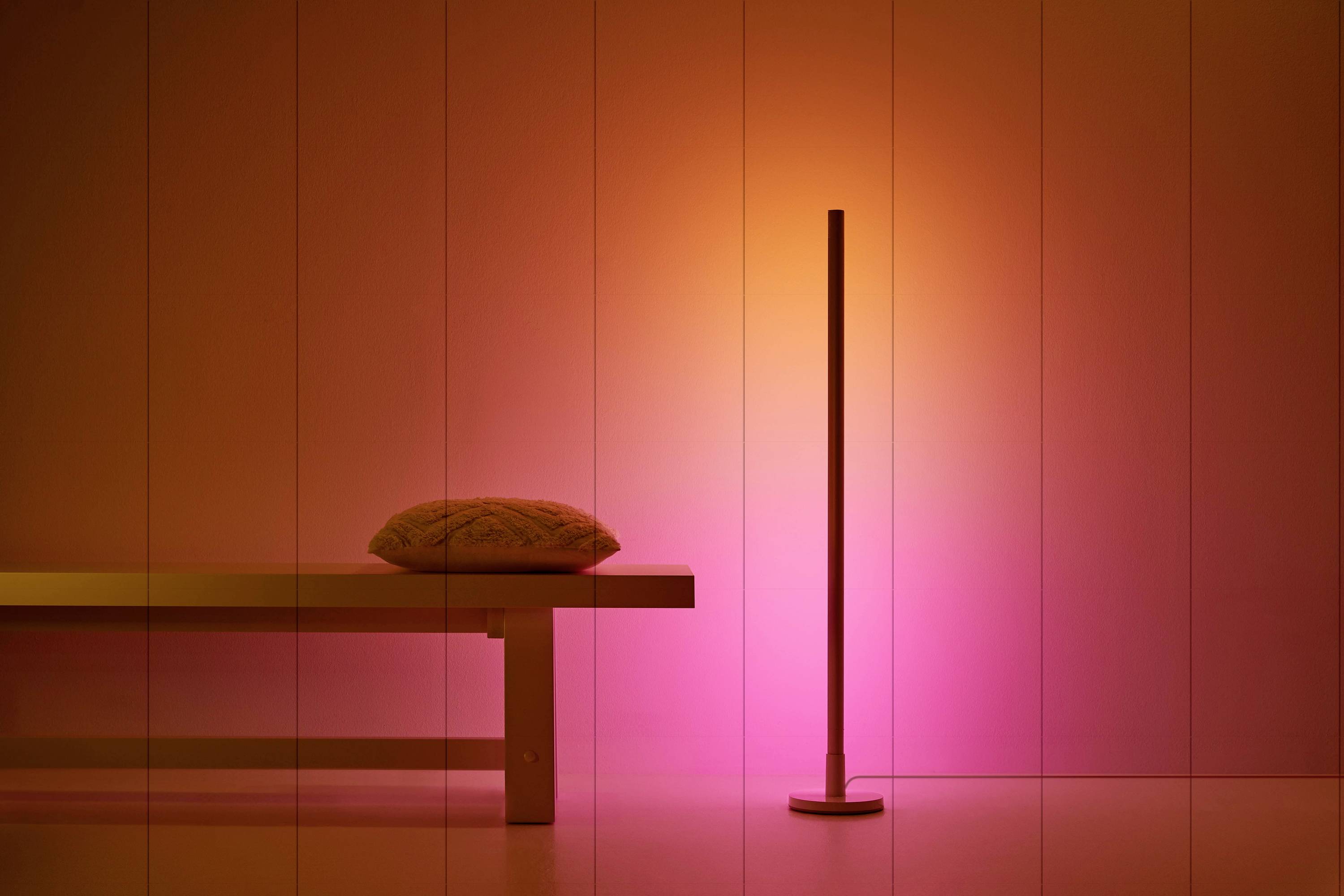 A bench with a cushion stands next to a slender, illuminated lamp. The background is lit in warm pink and orange hues.