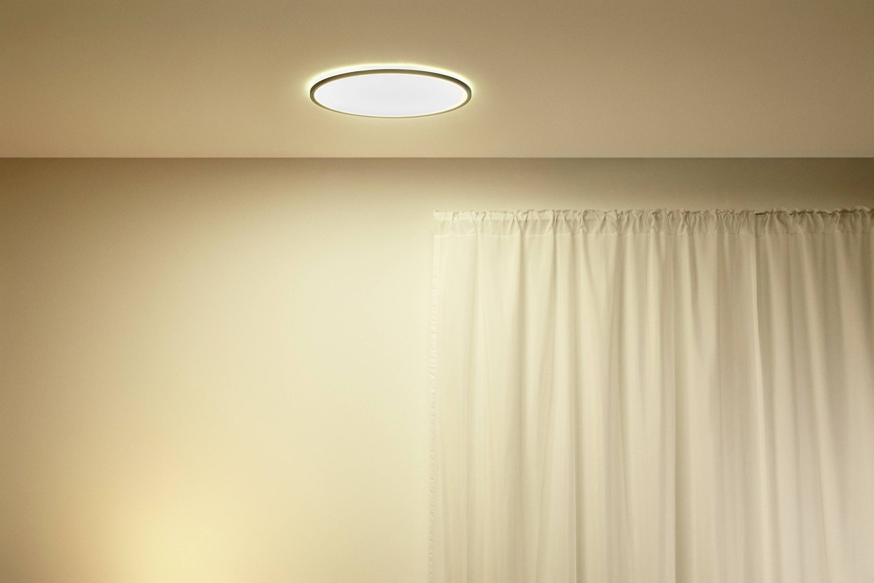 A modern, circular ceiling light emits warm light, illuminating a room with cream-coloured walls and a white curtain.