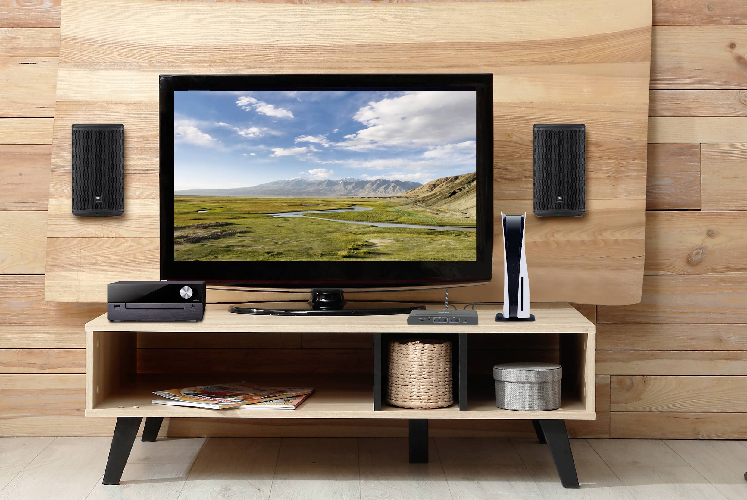 A television on a wooden shelf displays a landscape with mountains and sky. Beside it are speakers and gaming consoles.