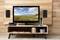 A television on a wooden shelf displays a landscape with mountains and sky. Beside it are speakers and gaming consoles.