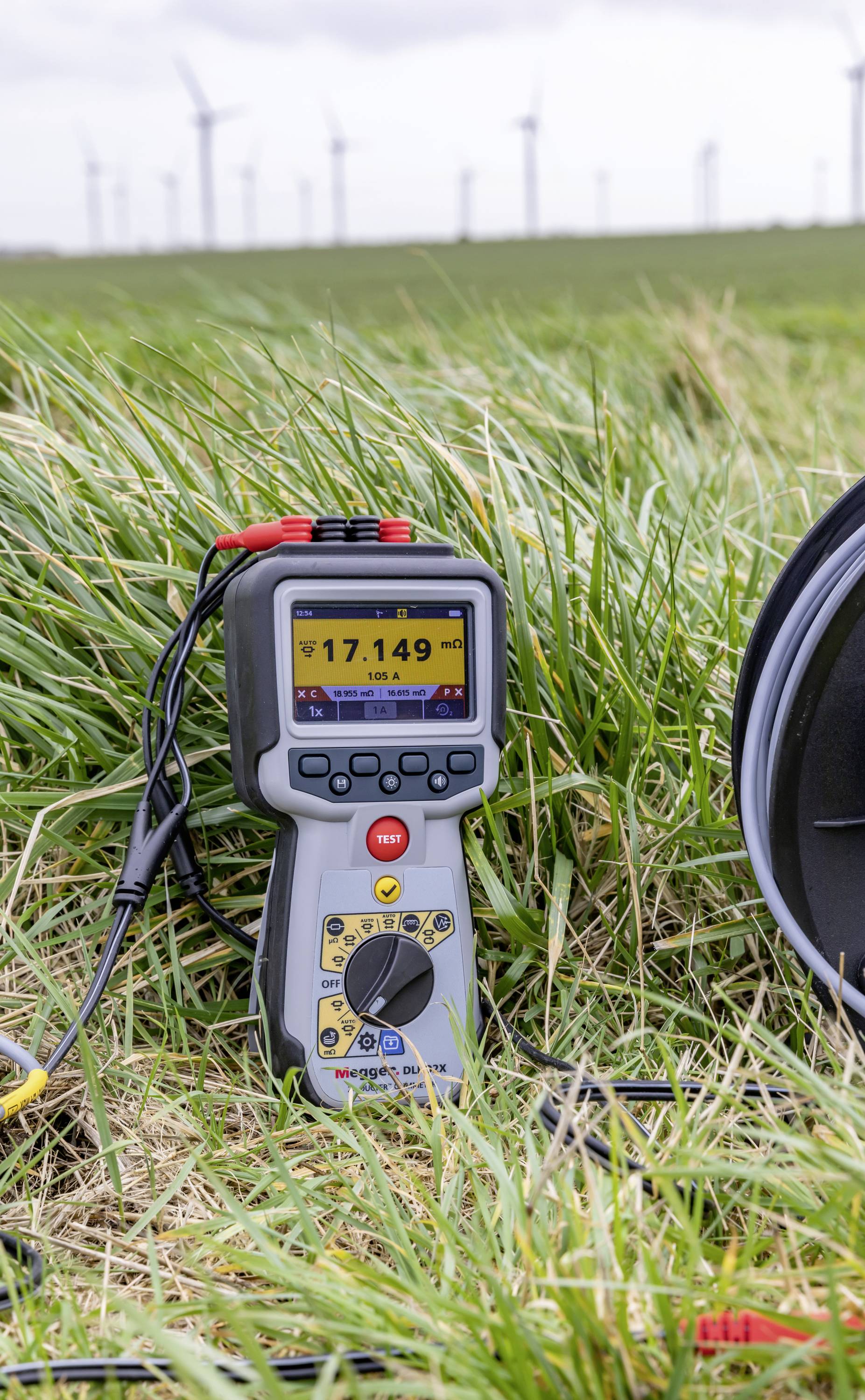 A multimeter lies in the tall grass in front of a wind farm. It displays '17.149' on the screen and is surrounded by cables.