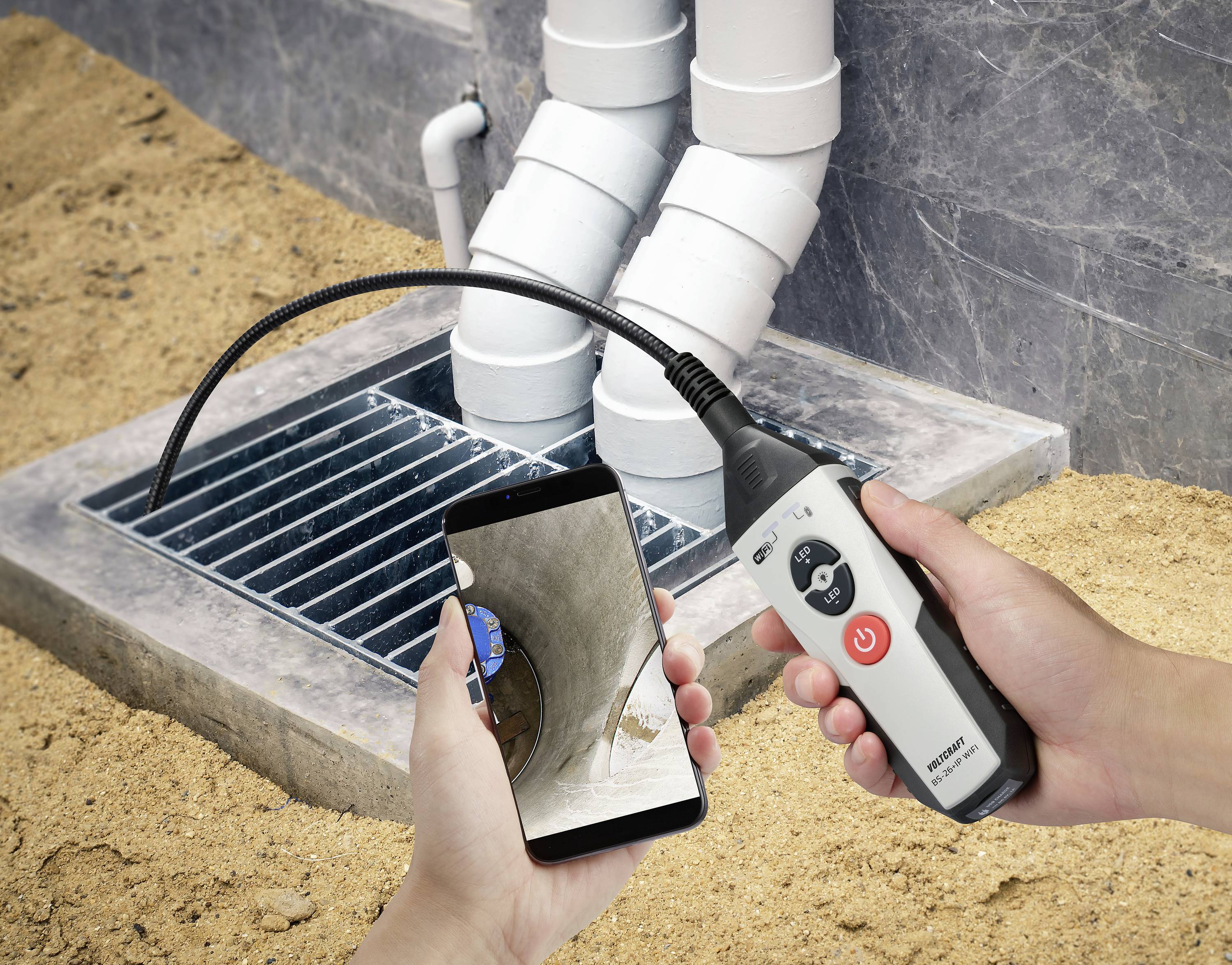 A person is holding an endoscope and a smartphone. The endoscope is examining a pipe within a drainage grid. The image shows its use for inspection.