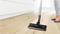 A person is hoovering a light wooden floor with a black vacuum cleaner. White kitchen cupboards are visible in the background.