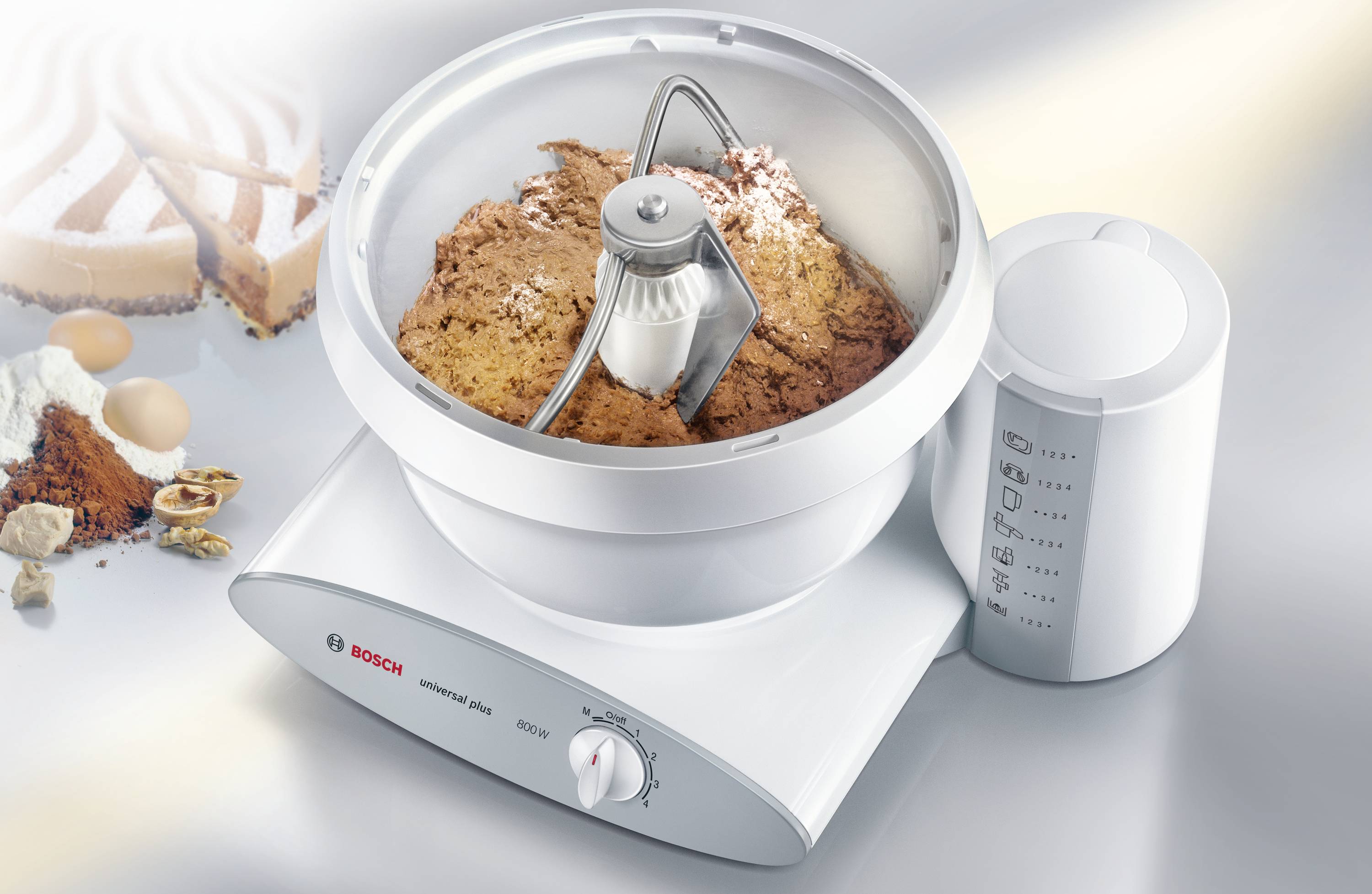 'Food mixer with dough hook kneads dough in a bowl. Beside the machine, ingredients such as eggs and flour can be seen.'