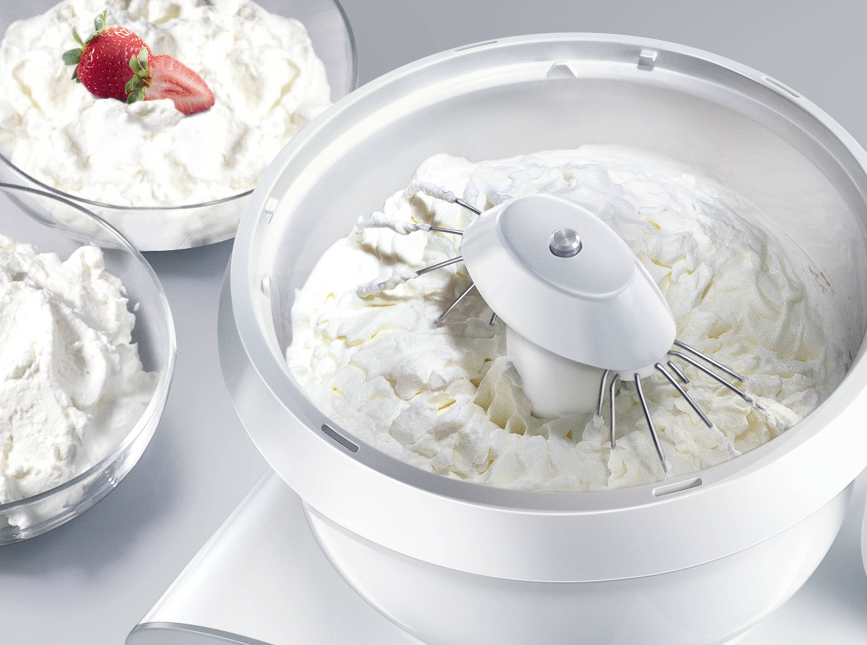 A food mixer is whisking cream until stiff. In the background, bowls of whipped cream can be seen, one garnished with strawberries.