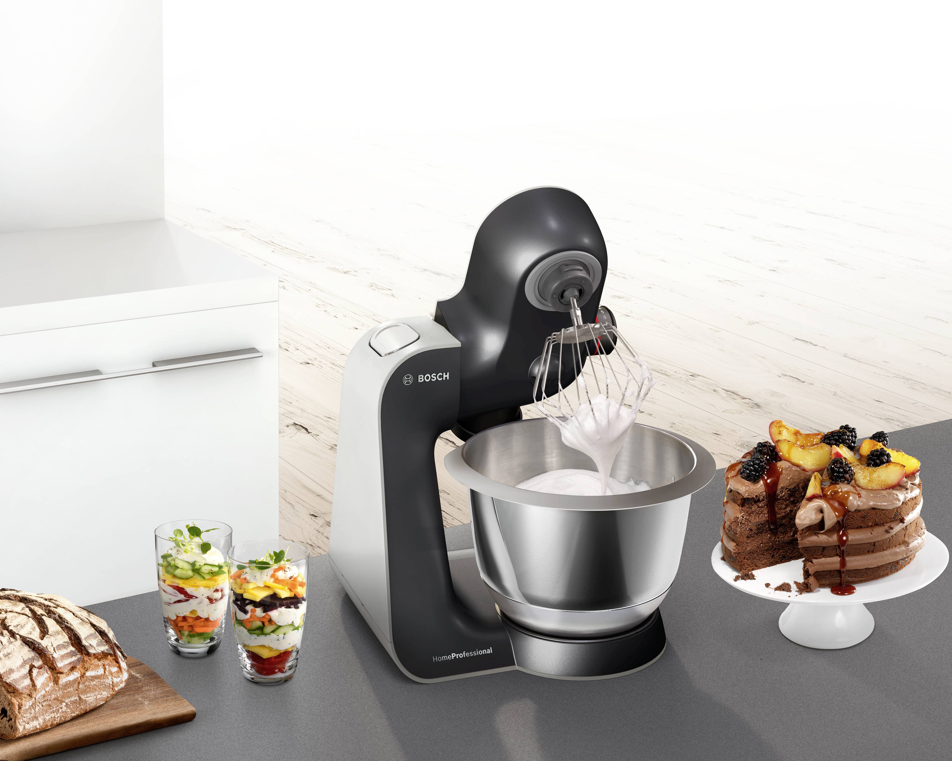 A food mixer is stirring a mixture in a metal bowl. Beside it, a cake, two dessert glasses and a loaf of bread sit on the worktop.