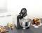 A food mixer is stirring a mixture in a metal bowl. Beside it, a cake, two dessert glasses and a loaf of bread sit on the worktop.