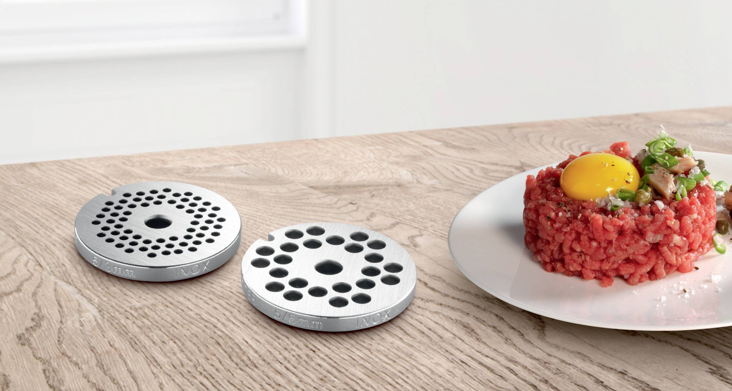 Two metal mincer blades on a wooden table next to a plate of steak tartare and a raw egg yolk as decoration.