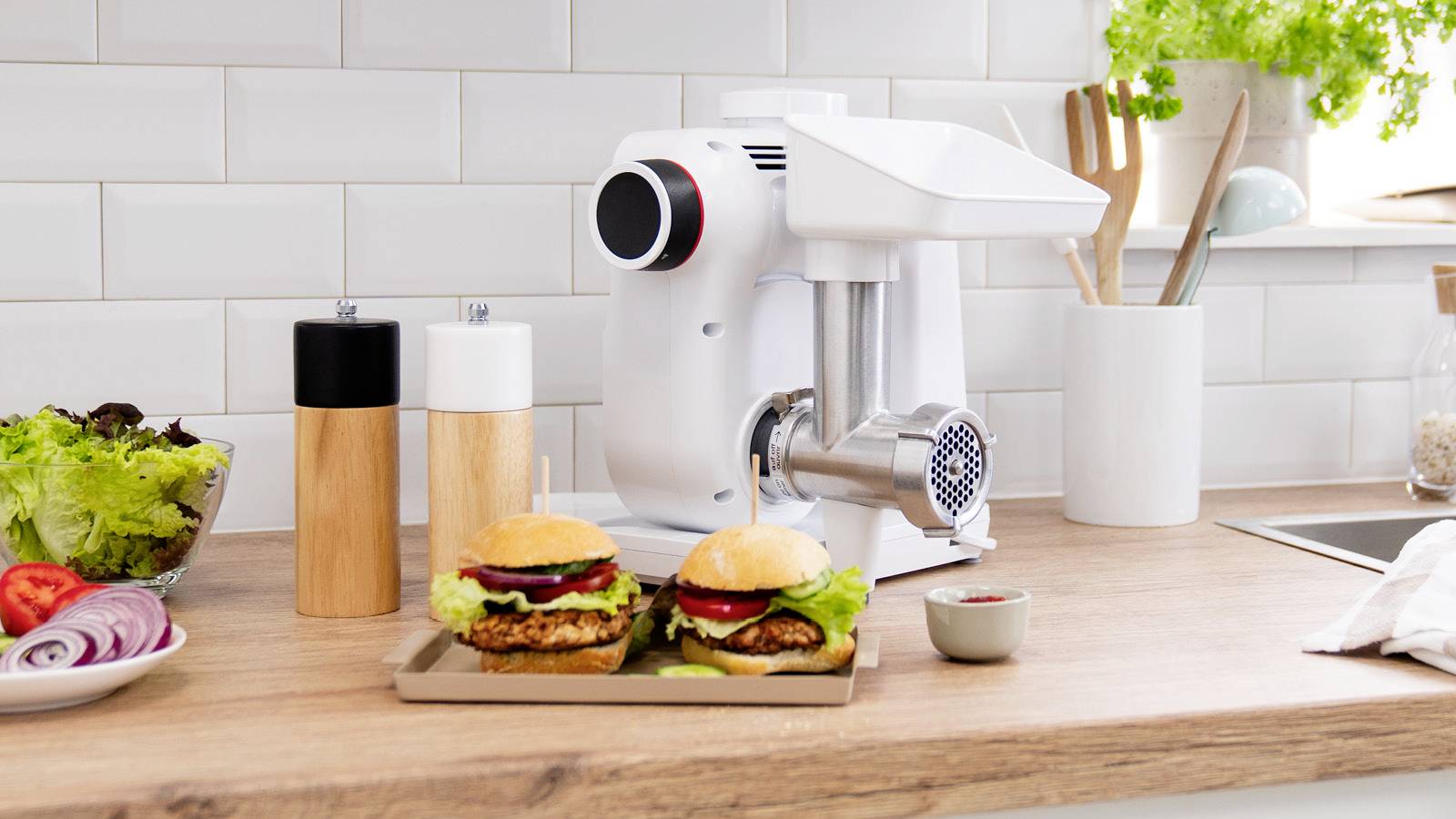 Two sandwiches with vegetables and meat in front of an electric mincer in a modern kitchen. Beside it are spice mills and salad.