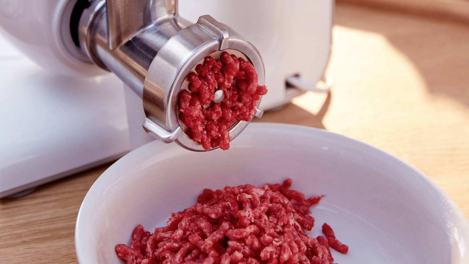 Fresh minced meat comes out of a mincing machine into a white bowl. The scene shows the process of meat preparation.