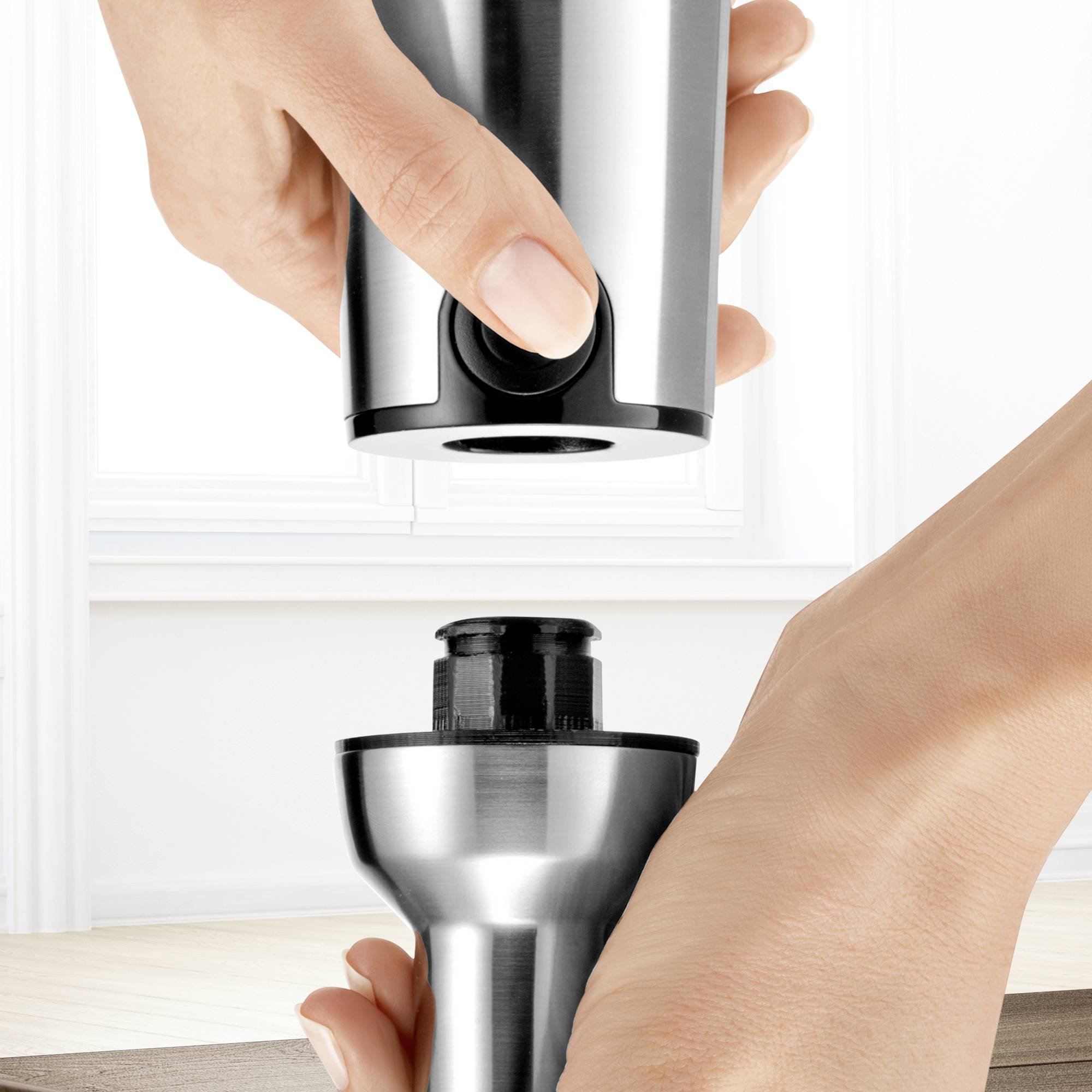 A hand is holding a metallic stick blender, which is standing on a wooden table, ready for use in a bright kitchen.