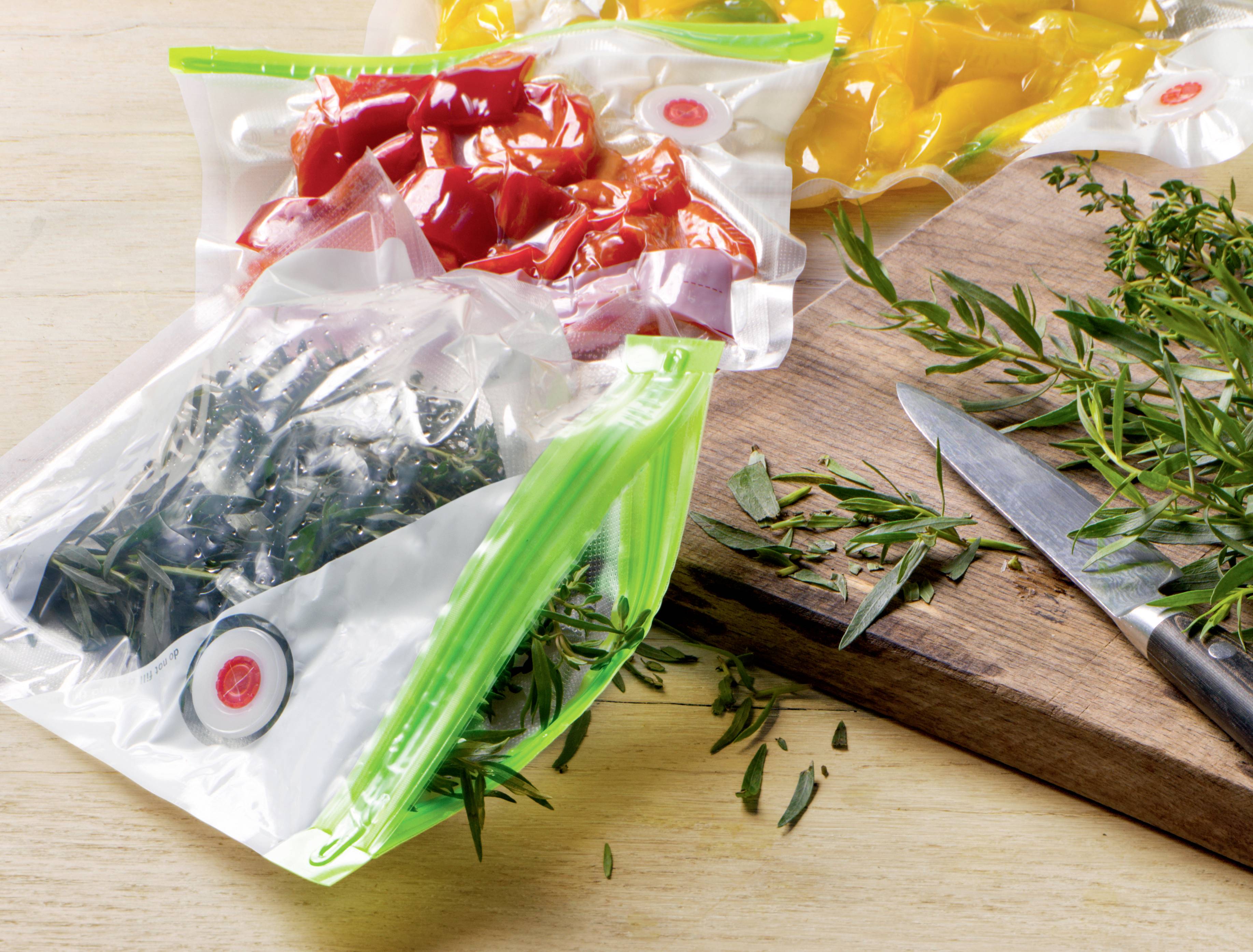 Three vacuum-sealed bags of coloured vegetables (red pepper, yellow pepper, green leafy vegetables) beside a knife on a wooden chopping board.