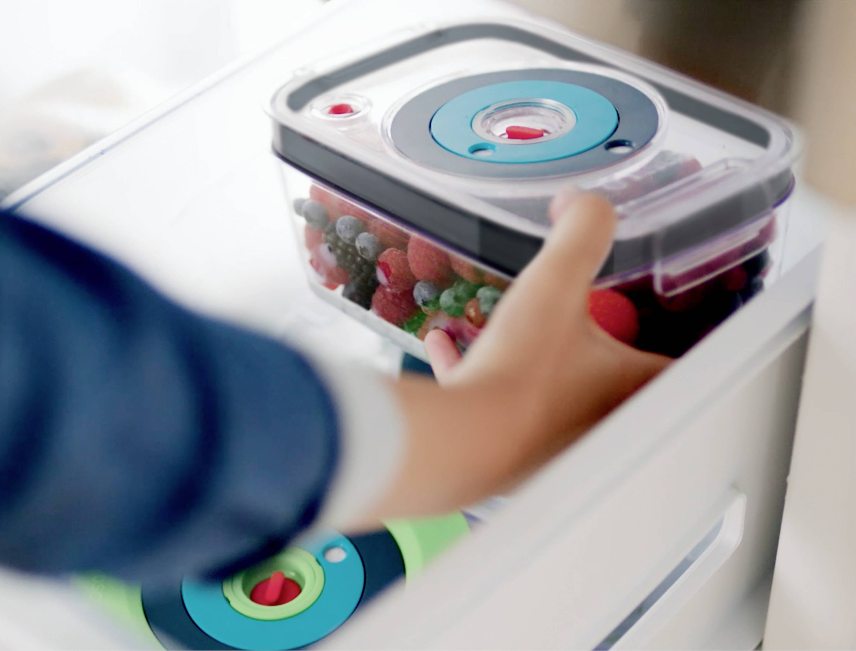 A hand places a sealed plastic container of berries into a fridge or freezer.
