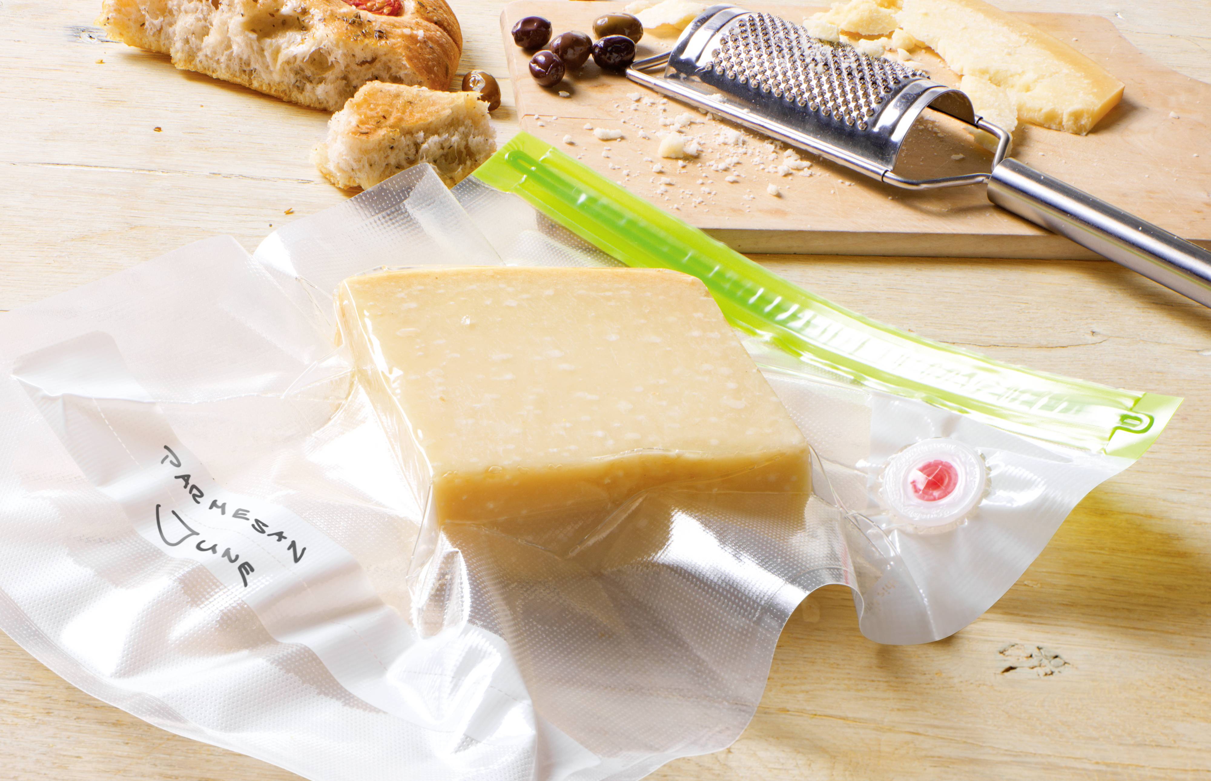 A piece of Parmesan in a vacuum-sealed bag lies on a wooden table, with a grater and bread beside it.