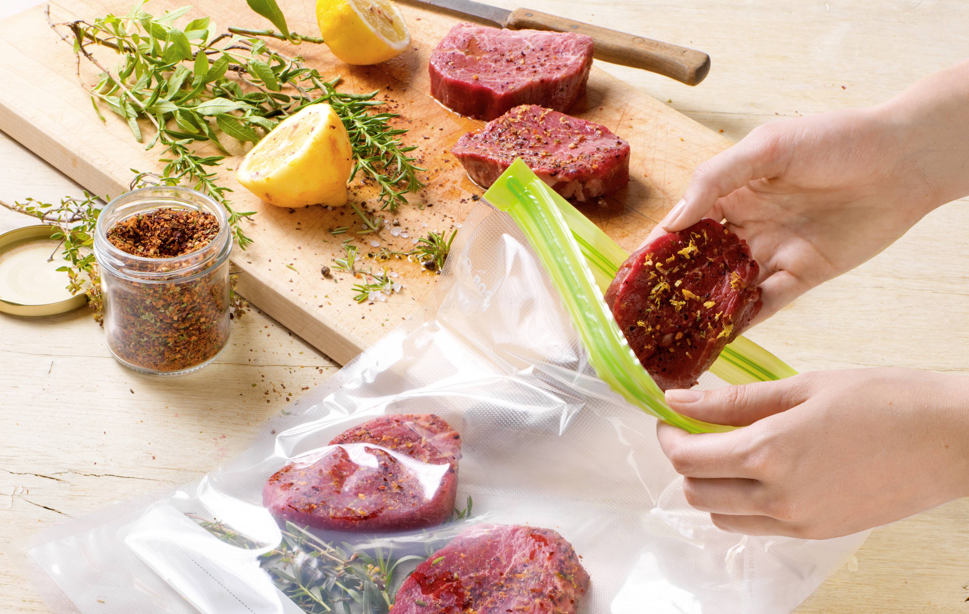 A person places seasoned meat into a sealable plastic bag; herbs, lemon, and spices are lying on a board.