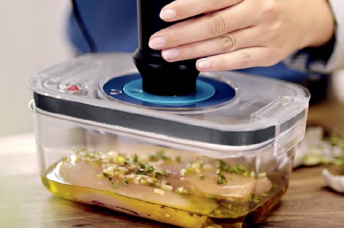 A person seals a plastic container with chicken breast and marinade using a vacuum sealer.