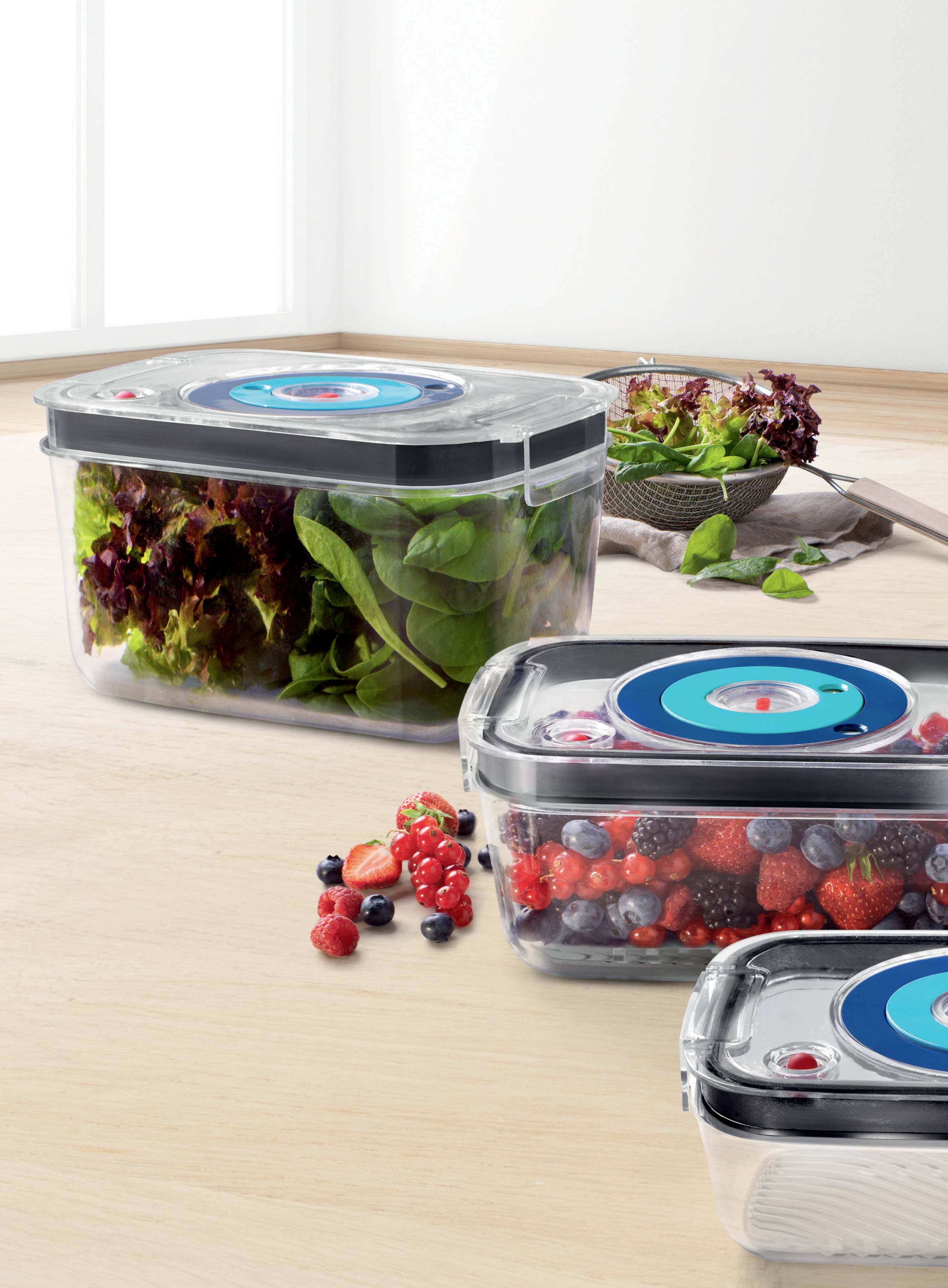 Two storage containers with preservation function are filled with berries and leafy salad. Alongside, additional fresh vegetables are placed.