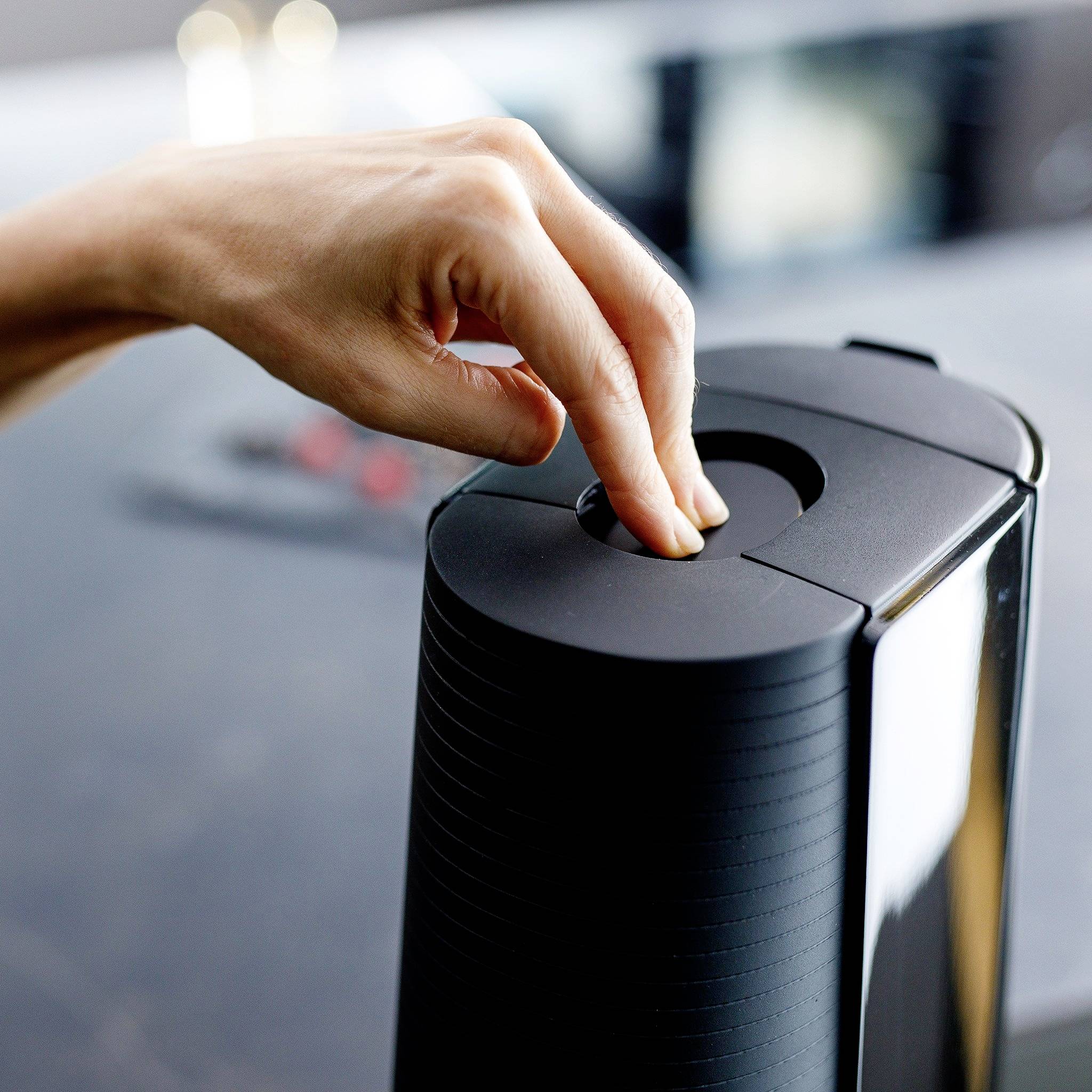 A hand presses the lid of a black electric kitchen waste bin. Background blurred.