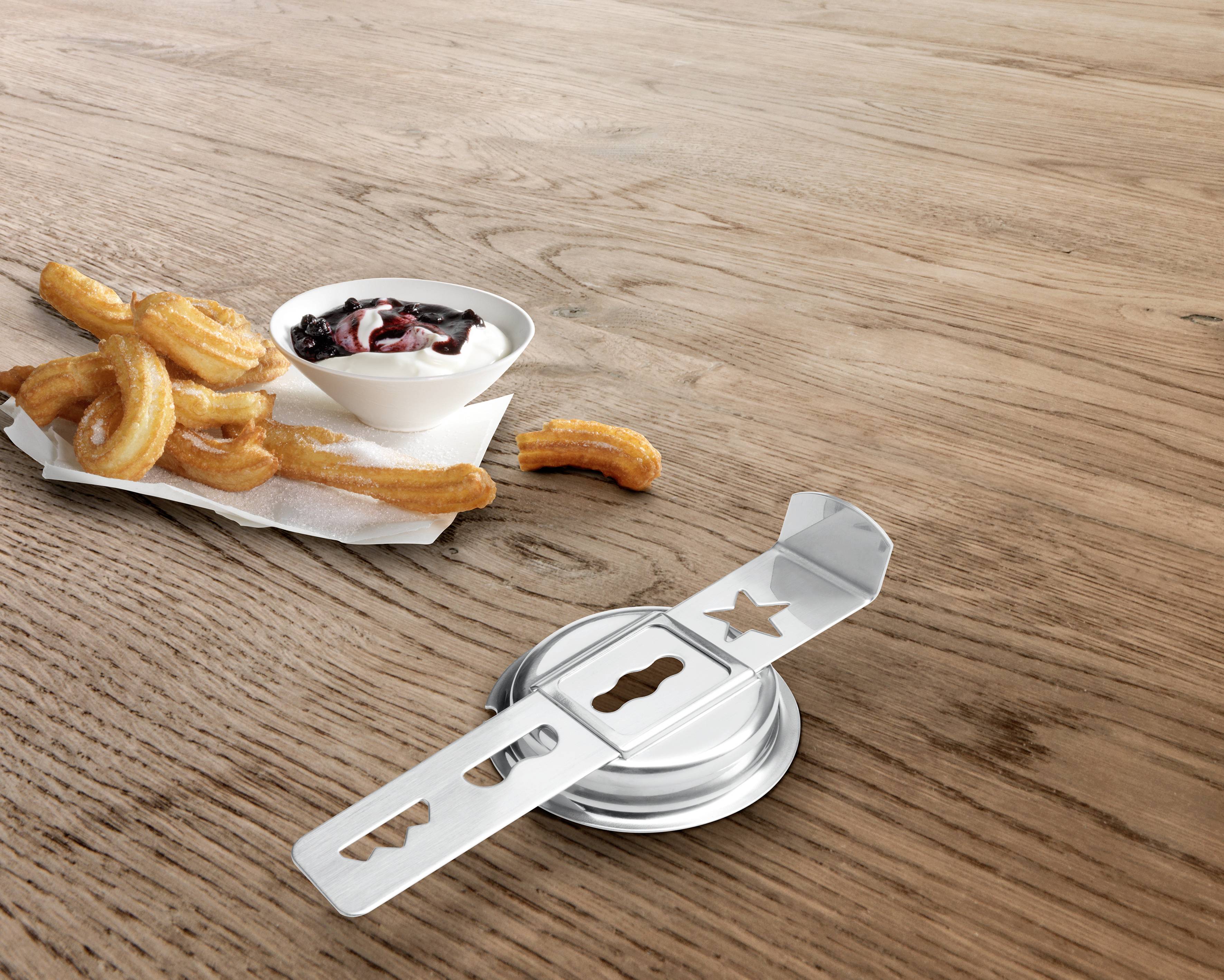 A metallic tin opener lies on a wooden table next to a bowl of churros and a small dish of sauce.
