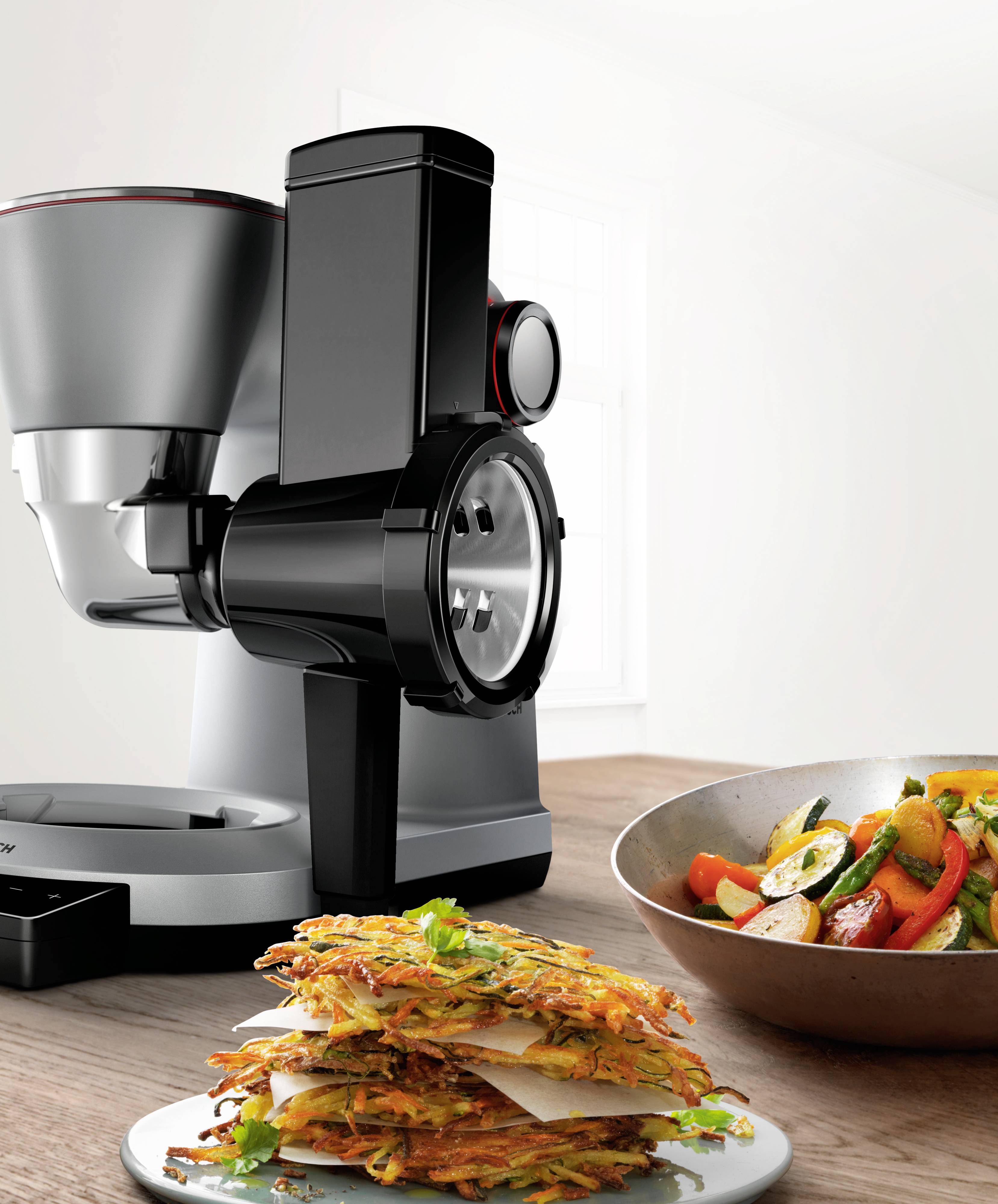 Food processor with a slicing attachment on a table, beside it a bowl of chopped vegetables and a plate of vegetable fritters.