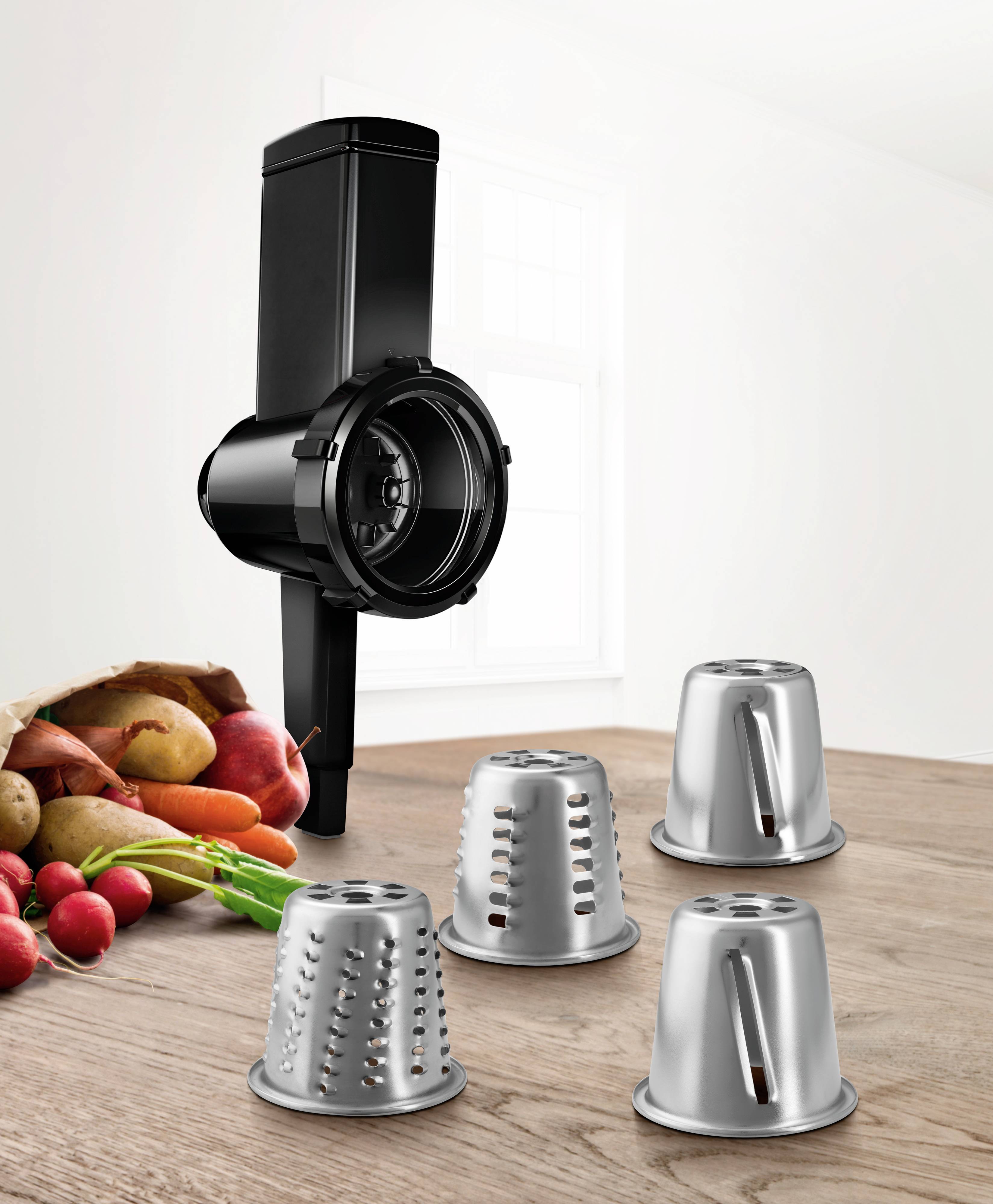 A black, glossy grater hangs on the wall. Next to it, four different drum attachments are placed on a wooden surface. In the background, fresh vegetables such as radishes and potatoes can be seen.