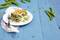 A plate with a portion of vegetable lasagne on a blue wooden table, surrounded by fresh pea pods. The plate is resting on a small fabric cloth.
