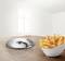 A white bowl of chips sits on a wooden table next to a flat, round silver device, with a window in the background.
