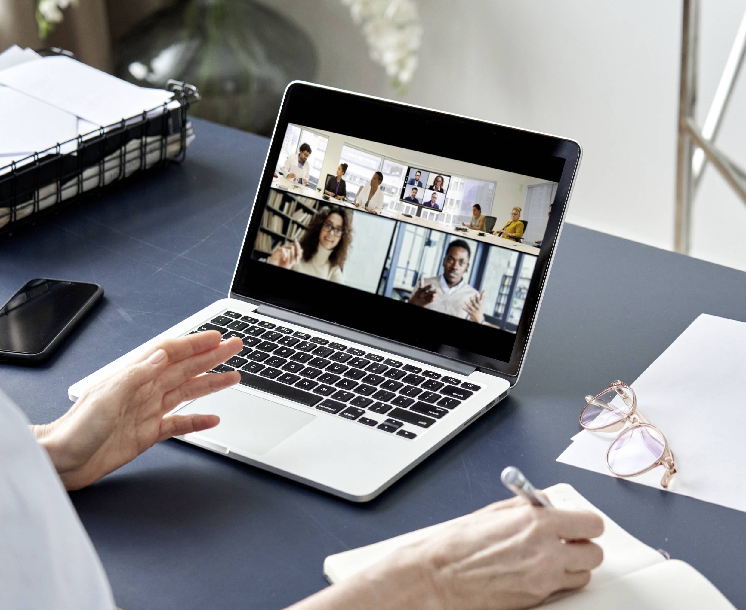 A person is participating in a virtual meeting on a laptop with multiple participants. Notes are being taken on a notepad.