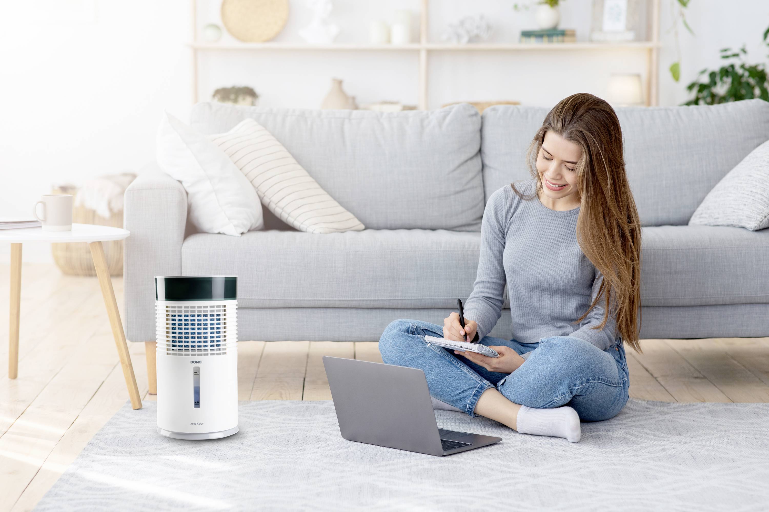 A woman is sitting on a carpet in a modern living room, writing in a notebook, with a laptop in front of her; an air purifier is next to her.