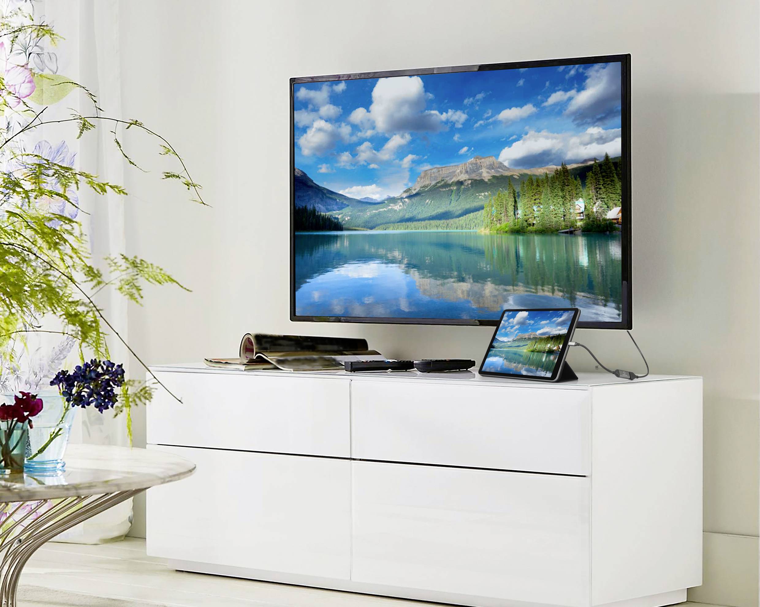 A television on a white cabinet displays a landscape with a lake and mountains. A tablet with a similar image stands beside it.
